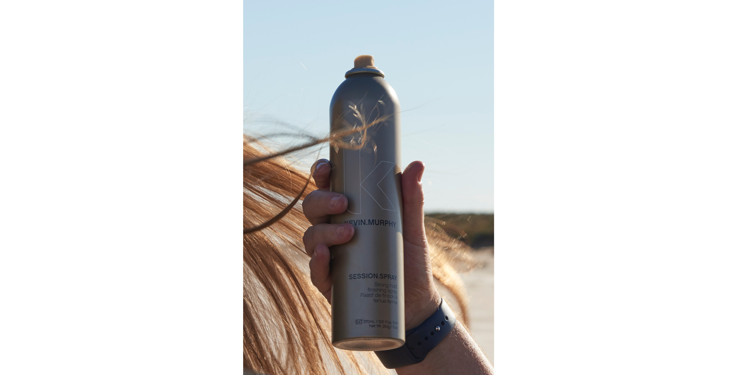 Hand holding a KEVIN.MURPHY SESSION.SPRAY against a windy beach backdrop.